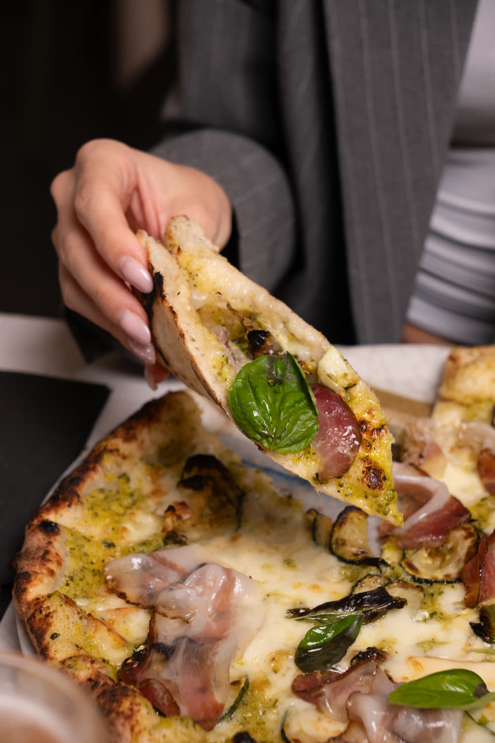 Una mano solleva uno spicchio di pizza con crema verde, salume e basilico